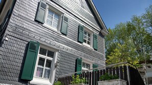 A two-story building with a grey shingled exterior, green shutters on the windows, and a gated entrance with decorative planters.