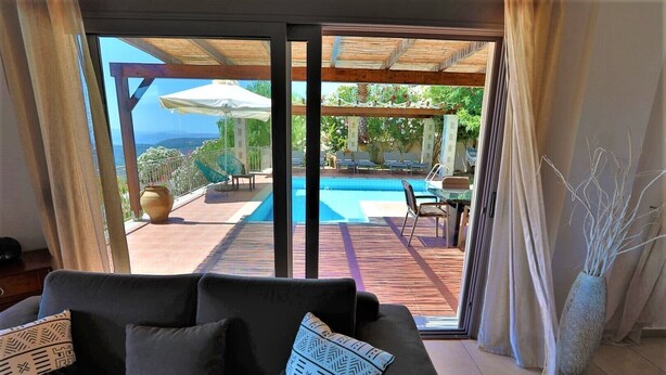 Ett rum med en stor skjutdörr i glas som leder ut till en uteplats med en pool, en möblerad sittgrupp och utsikt över havet.