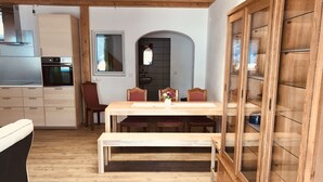 A dining area with a wooden table, chairs, and a bench. There is a wooden cabinet with glass doors to the right. A kitchen with cabinets and a window is visible in the background. - Holiday tower Niederstetten (Niederstetten)