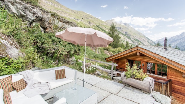 A mountain lodge with a glass table and white sofa, surrounded by greenery and a wooden cabin.