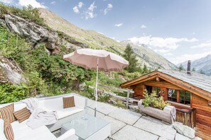 A mountain lodge with a glass table and white sofa, surrounded by greenery and a wooden cabin. - Single family house with private outdoor jacuzzi & sauna, ski in - ski out (Zermatt)