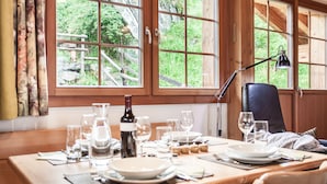 A dining table set with plates, glasses, and a wine bottle, with a view of greenery outside through large windows.