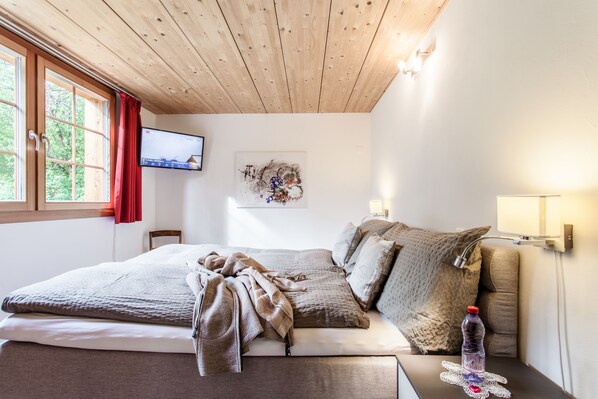 Un dormitorio con techo de madera, cama con manta, almohada, botella de agua, televisor en la pared y ventana con cortinas rojas.