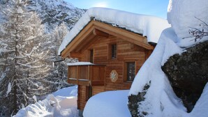 A wooden cabin with a snow-covered roof, surrounded by snow-covered trees and mountains.