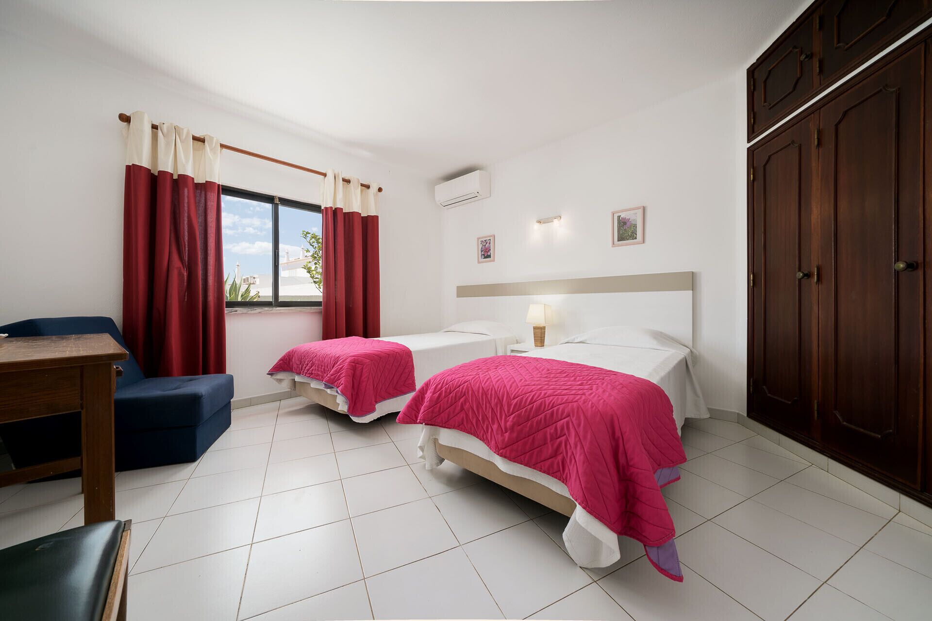 A hotel room with two beds, a desk, a chair, a wardrobe, and a window with curtains.