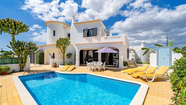 Una villa blanca con piscina, estructura de dos plantas y un patio con sillones de descanso.