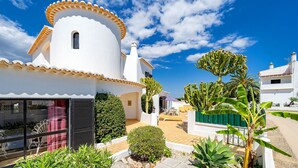 Una villa blanca con techo curvo, un balcón pequeño y un jardín con cactus y palmeras.