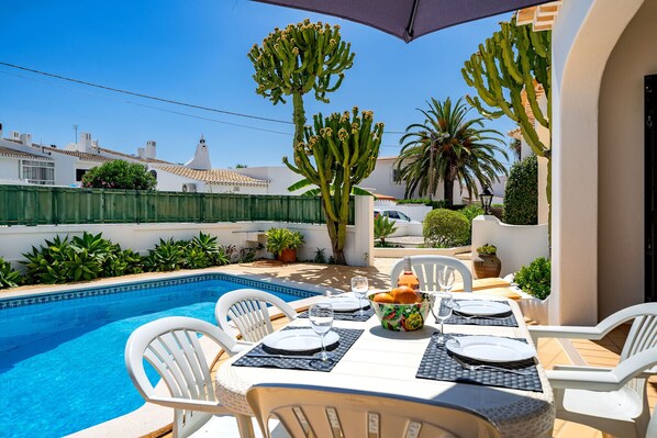 Un área de comedor junto a la piscina con una mesa preparada para cuatro personas, rodeada de sillas blancas, una piscina y cactus.