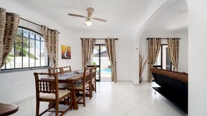 Un comedor con una mesa y sillas de madera, un ventilador de techo y vista a una piscina por las ventanas.
