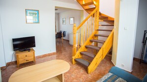 Una sala de estar con una escalera de madera, un televisor de pantalla plana sobre un mueble de madera y un comedor al fondo.