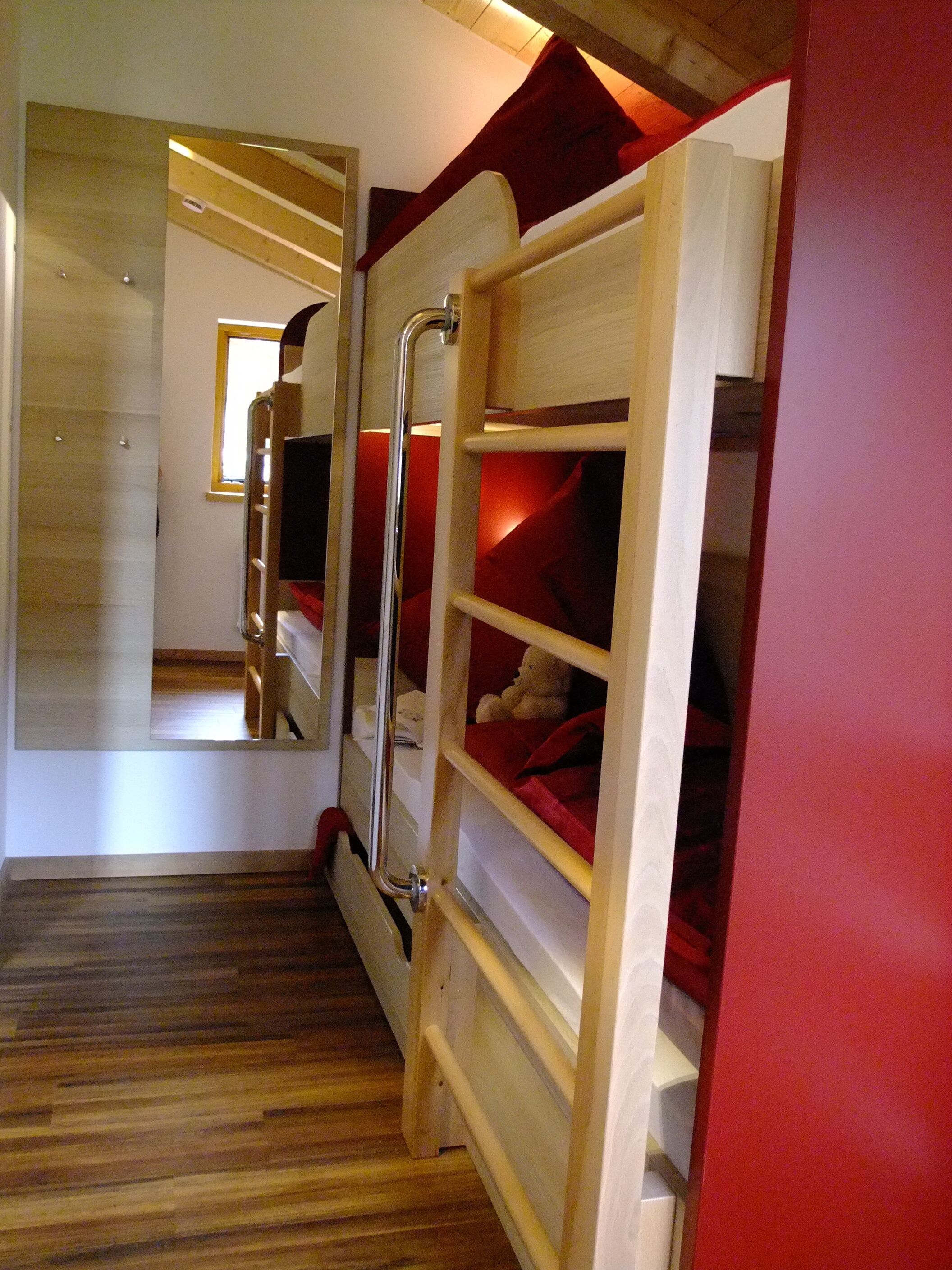 Een stapelbed met een rode hoofdbord, houten vloer en een spiegel die de kamer weerkaatst.