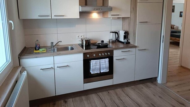 Une cuisine compacte avec des meubles blancs, un évier en acier inoxydable, une cuisinière et une bouilloire.