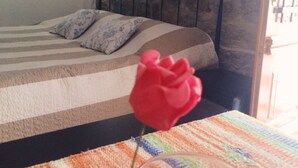 Uma cama com colcha listrada e duas almofadas, uma rosa vermelha em vaso de vidro sobre toalha de mesa listrada, e uma parede de pedra ao fundo da cama.