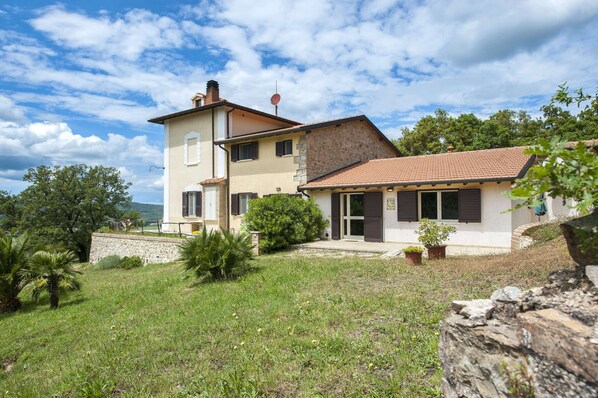 Une maison à deux étages, dotée d’une façade en pierre et d’un toit recouvert de tuiles, entourée d’une cour verdoyante et d’arbres.