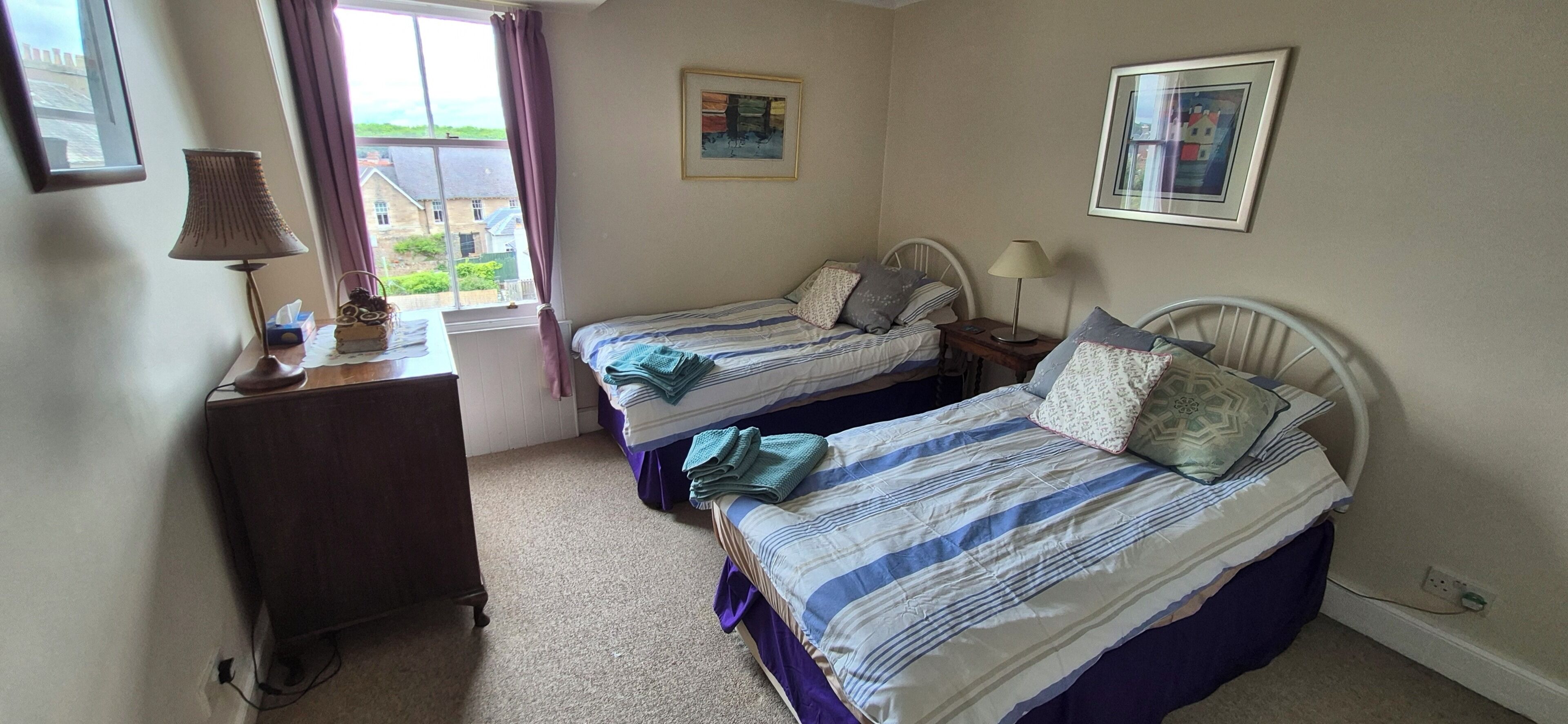 Een slaapkamer met twee eenpersoonsbedden, een nachtkastje met een lamp, een dressoir en een raam met gordijnen.