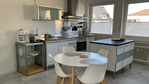 Cozinha moderna com armários brancos, mesa de jantar com cadeiras e vista para o exterior através de grandes janelas.