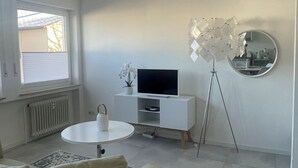 Uma sala de estar moderna com um sofá, uma pequena mesa redonda, uma TV sobre um móvel branco, um abajur e uma janela com persianas.