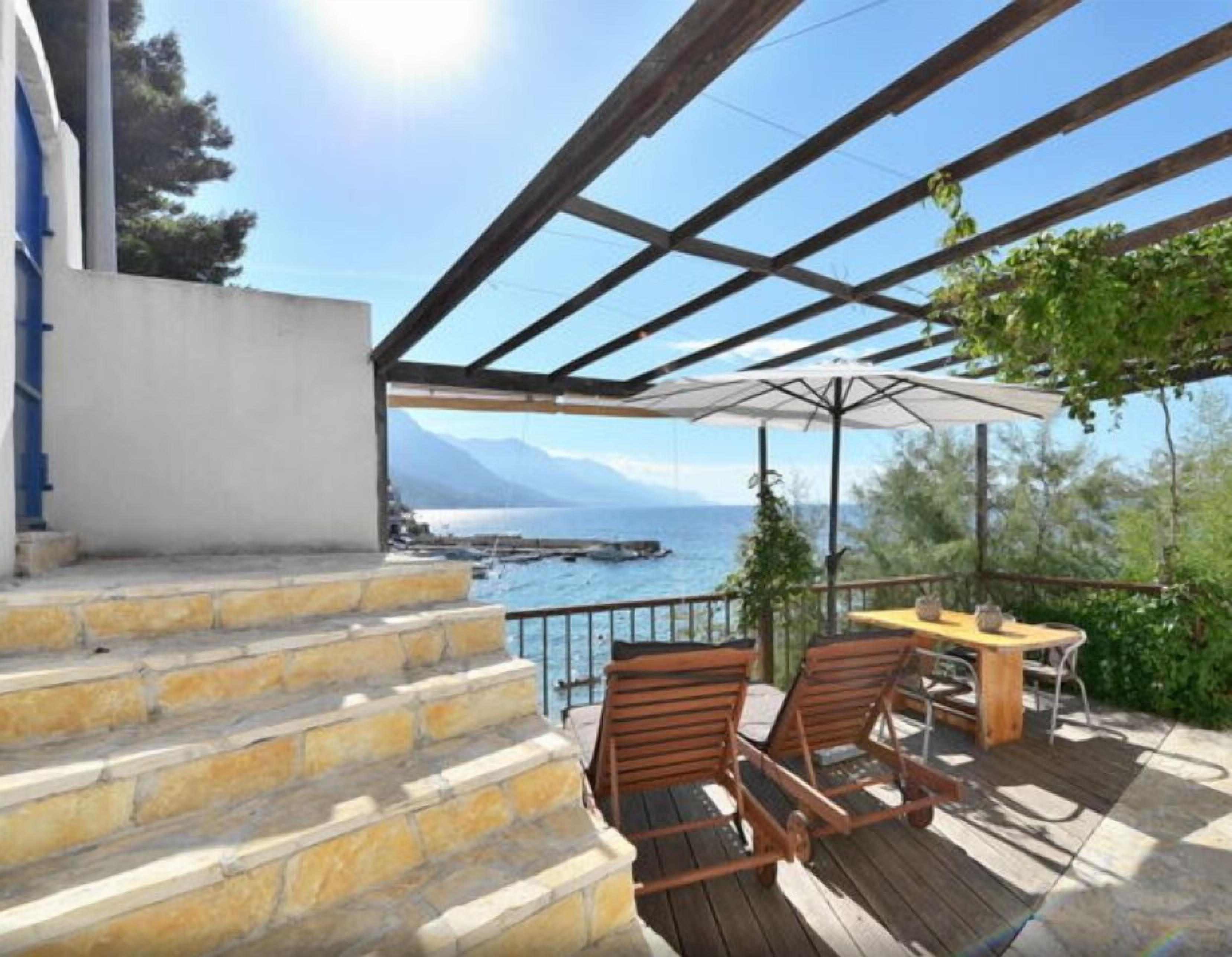A wooden deck with a table and chairs, overlooking a body of water and mountains.