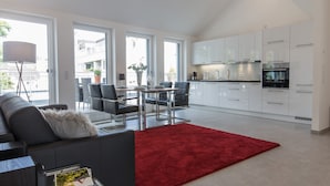 A modern kitchen with white cabinets and stainless steel appliances, a dining area with a red rug, and a balcony with a view of the city.