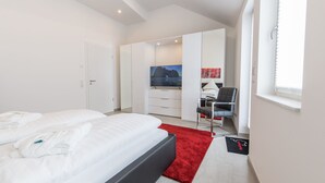 A modern hotel room with a large bed, a desk with a chair, a TV, and a view of the sea.