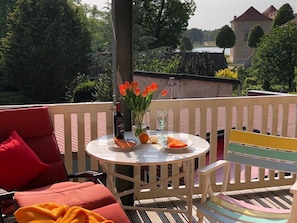 A round table with a bottle, glasses, and fruit on a balcony with a view of trees and a building.
