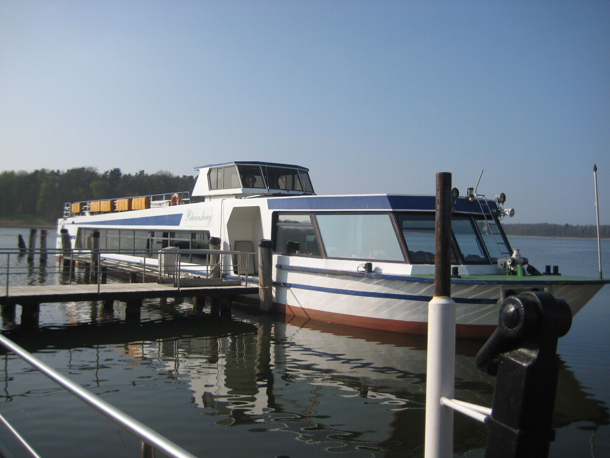 A boat docked at a pier with a clear view of the water and a forested shoreline in the background.