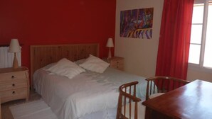 Een slaapkamer met een houten bed, een nachtkastje, een stoel, een tafel en een raam met rode gordijnen.