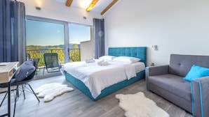A bedroom with a large bed, a grey sofa, a desk, and a balcony with a view of the mountains.
