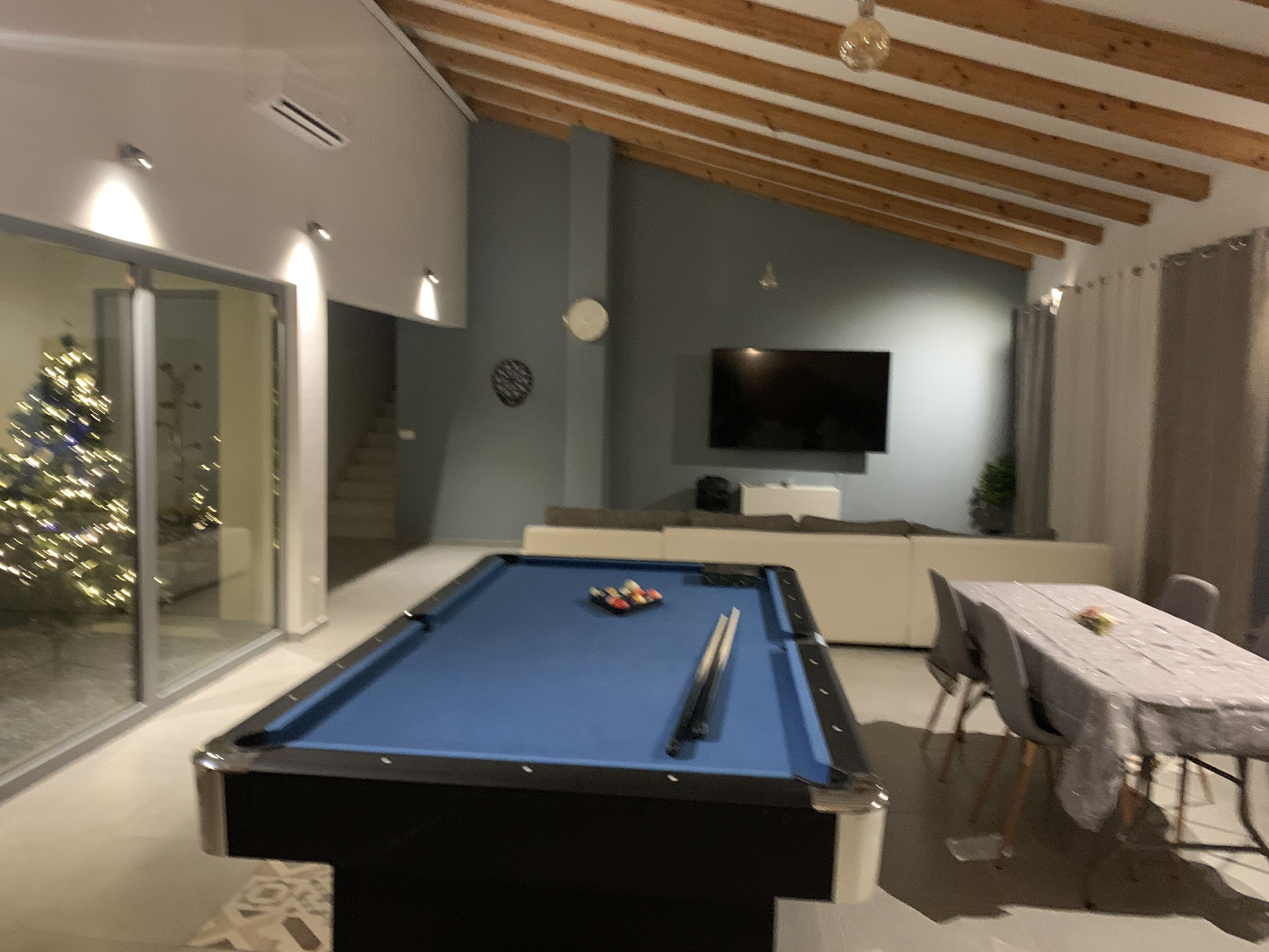 A pool table in a room with a TV mounted on the wall, a Christmas tree, and a dining table with chairs.