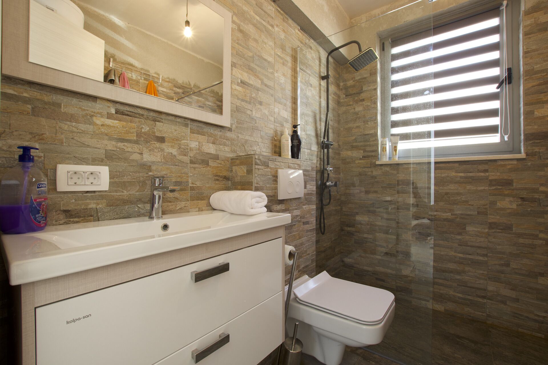 Een moderne badkamer met een achterwand van steen, een witte wastafel, een spiegel, een toilet en een douche met een glazen deur.