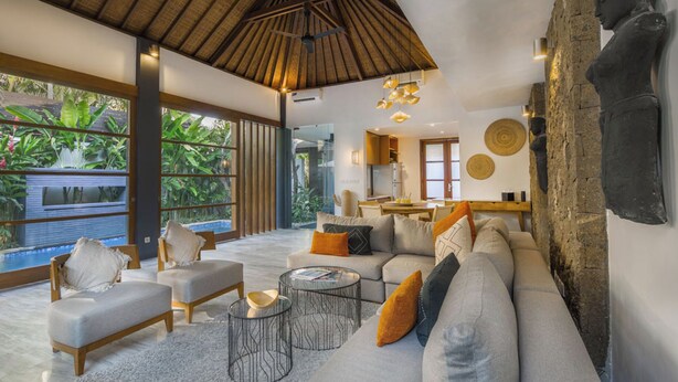 A spacious living area with a large sofa, a coffee table, and a dining table. There is a view of a swimming pool through large windows.