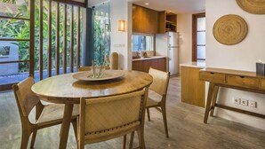 A dining area with a round wooden table and chairs, a kitchen in the background, and a view of greenery outside.