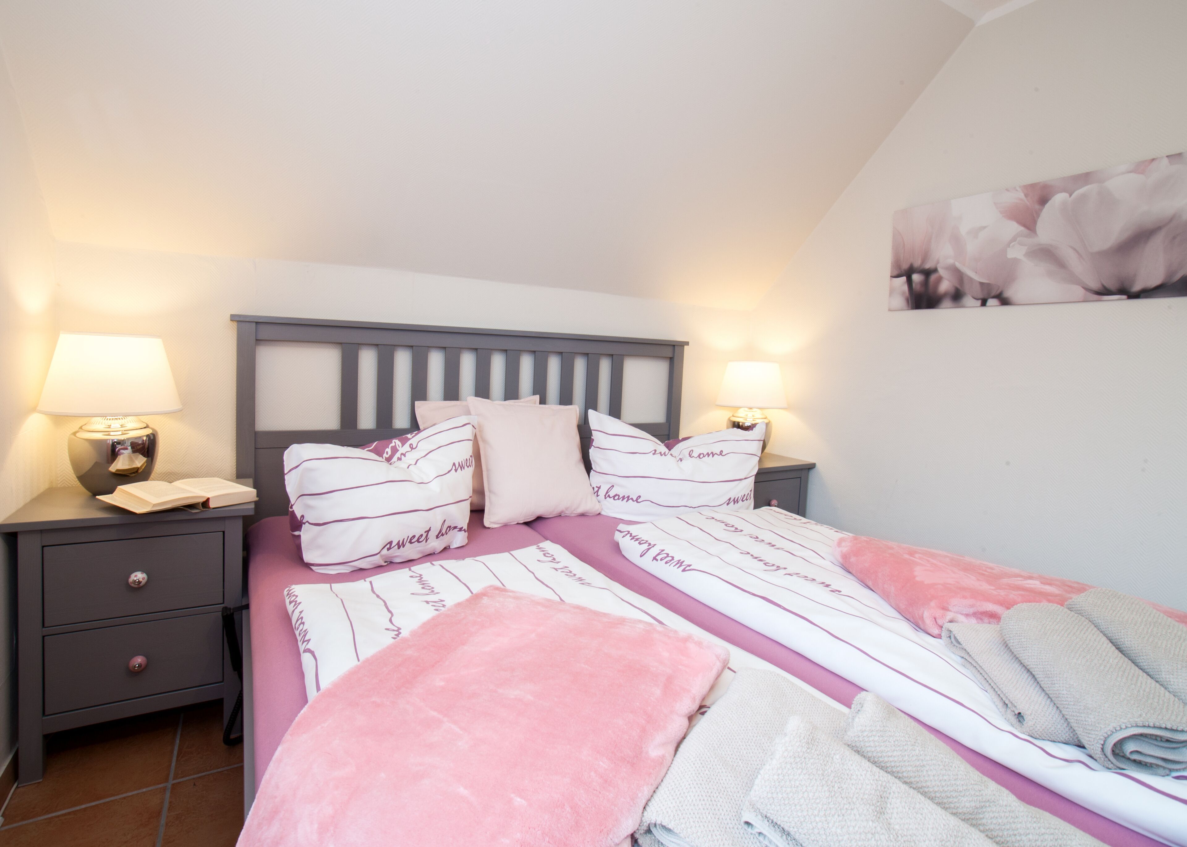 A bedroom with a bed, two bedside tables with lamps, a nightstand with a drawer, and a wall with a floral painting.