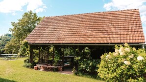 A rustic building with a tiled roof, a covered seating area with benches, and lush greenery surrounding it.