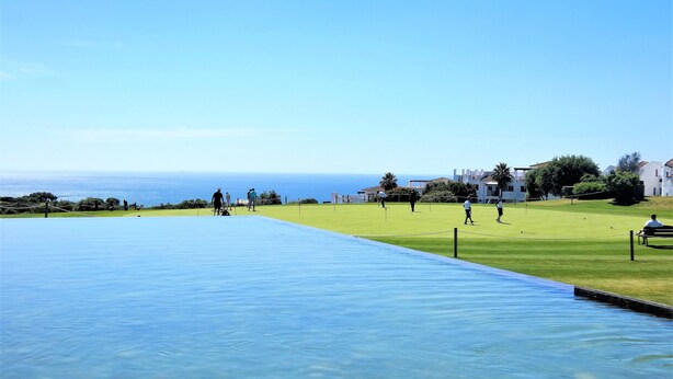 Una piscina con vista despejada al océano, un área verde con gente y edificios blancos al fondo.