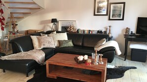 Una sala de estar con un sofá de cuero negro, una mesa de centro de madera y una alfombra blanco y negro. Hay cuadros enmarcados en la pared, una lámpara y una estantería llena de libros.