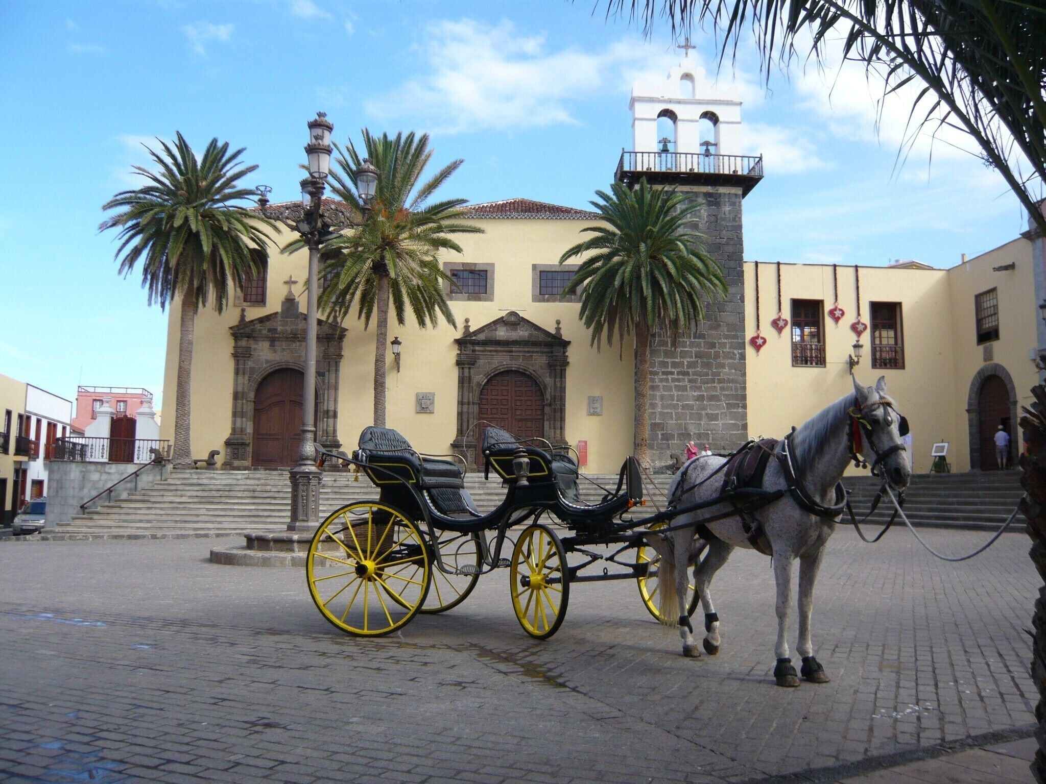 Un carruaje tirado por caballos estacionado frente a un edificio histórico con una torre de campanas y palmeras.