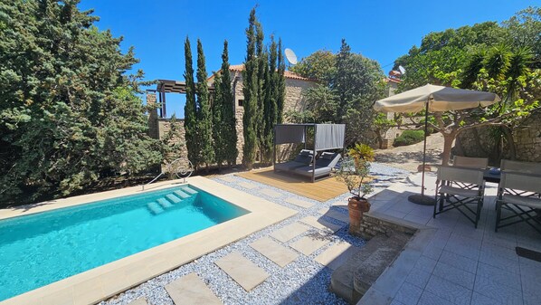 Una piscina con plataforma de madera, zona de estar cubierta y pared de piedra.
