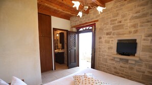 Un dormitorio con pared de piedra, techo de madera, cama con ropa blanca, televisor sobre una estantería de piedra y baño visible a través de una puerta abierta.