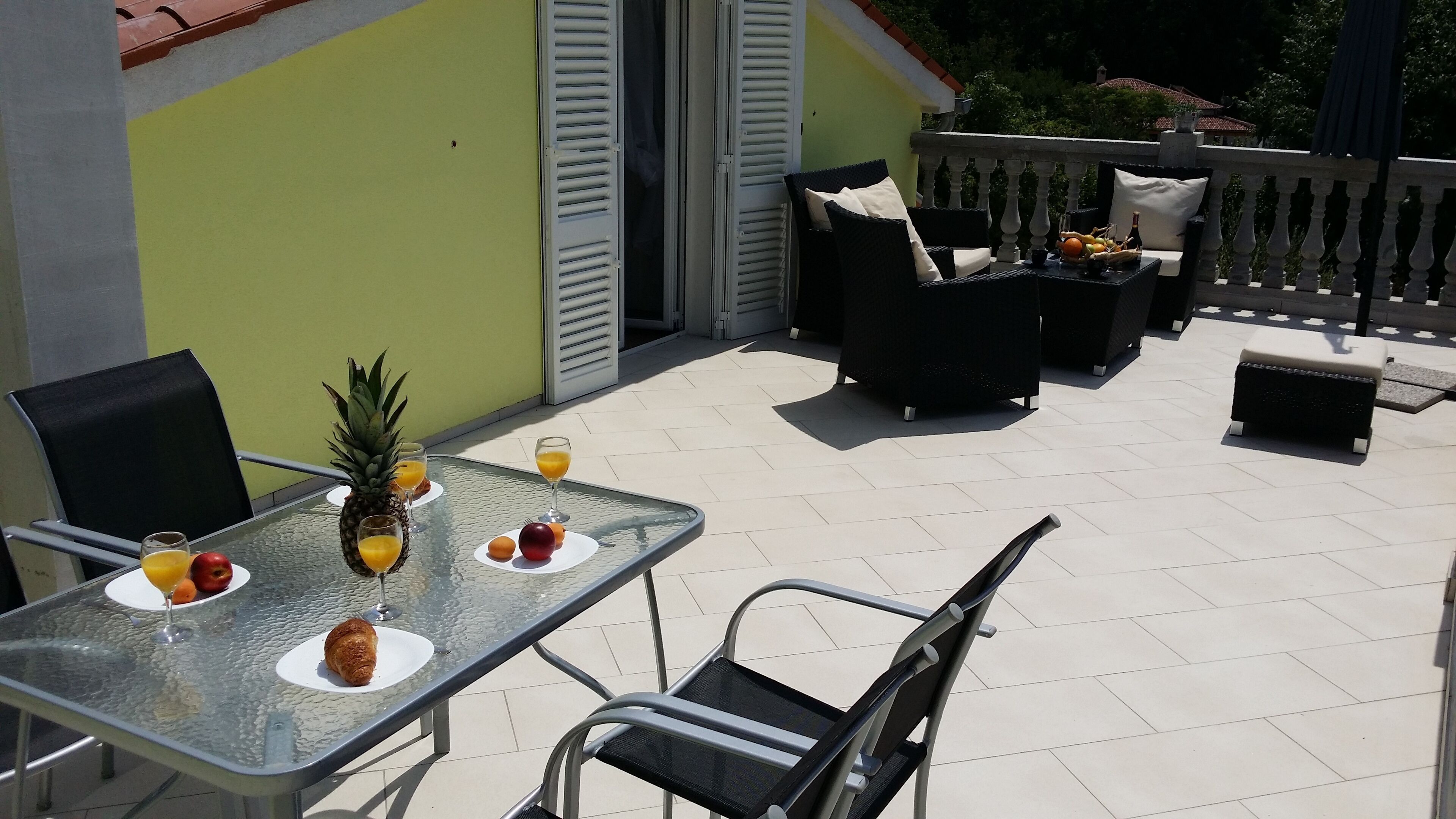 An outdoor patio with a glass table set for breakfast, including fruits and drinks.