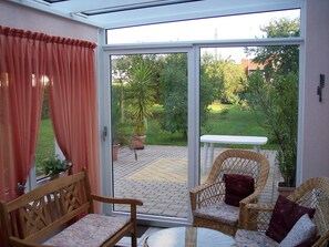 A room with a sliding glass door leading to a patio. There is a wooden chair with a cushion, a wicker chair with a cushion, and a small table. - Charming cottage with conservatory in Bayreuth on the eastern outskirts (Bayreuth)