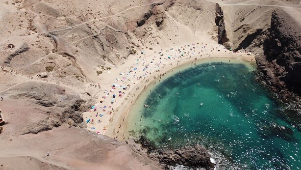 Une vue aérienne d’une plage aux eaux turquoise et limpides, avec un terrain rocheux et de nombreux parasols colorés.