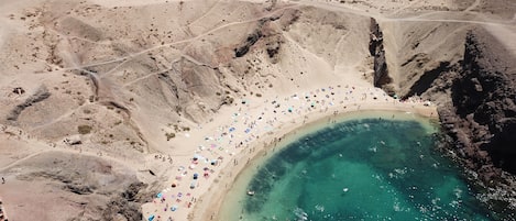Vista aérea de una playa con agua turquesa cristalina, terreno rocoso y numerosos sombrillas coloridas.