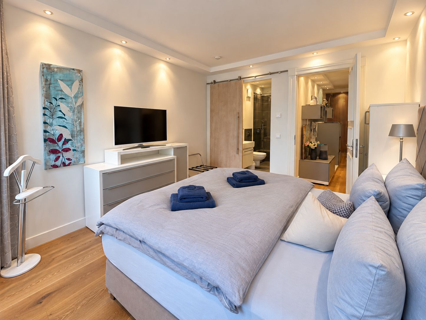 A modern bedroom with a bed, a flat-screen TV, a painting, and a bathroom visible through an open door.
