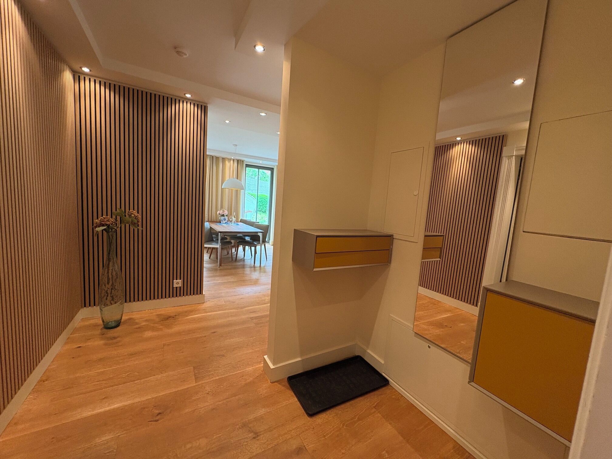 A hallway with wooden flooring, a black mat at the entrance, a vase with flowers, and a dining area in the background.