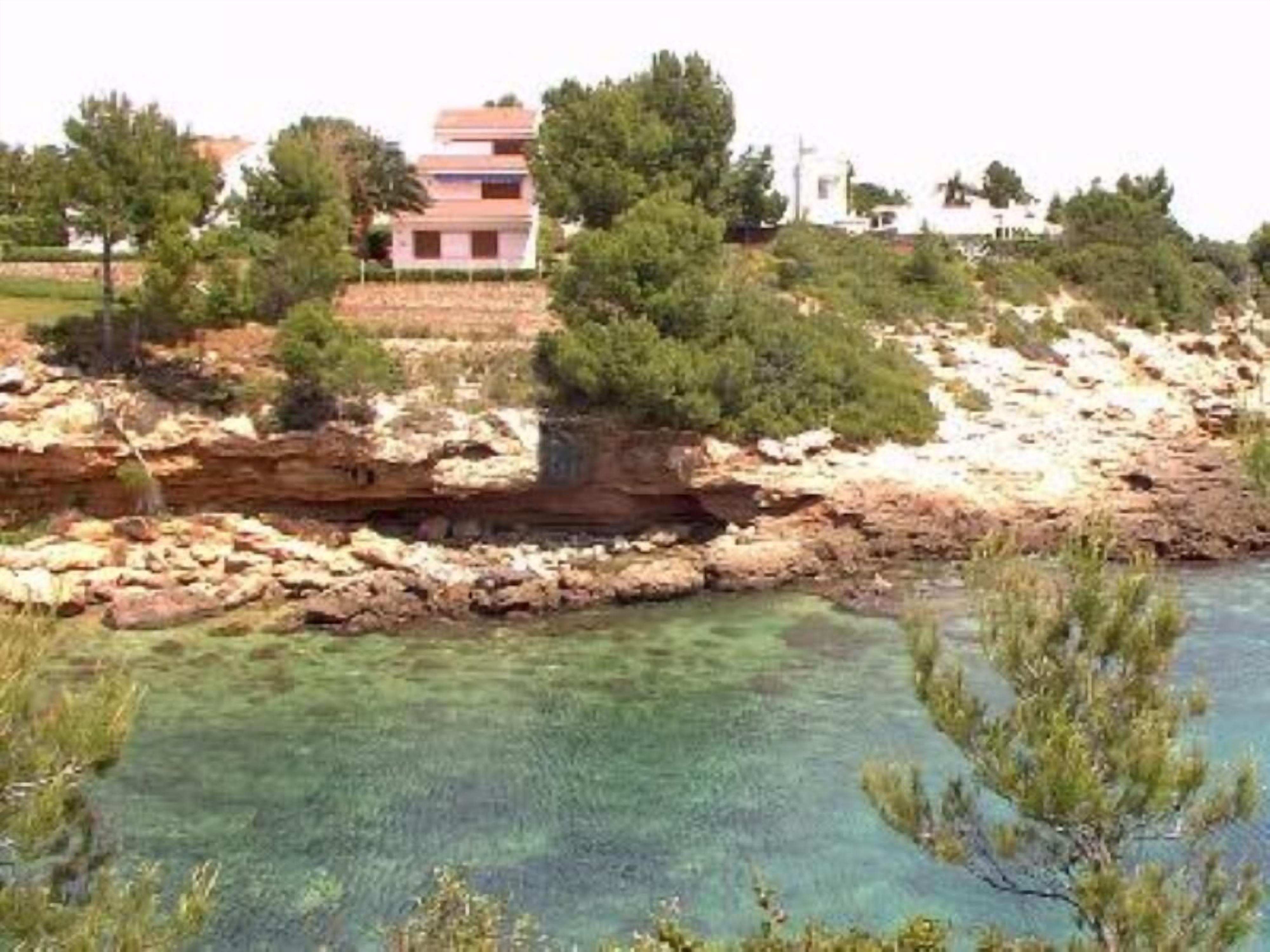 A coastal area with clear water, rocky terrain, and buildings in the background.