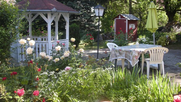 A gazebo with a white railing, a red shed, and a table with chairs in a garden setting.