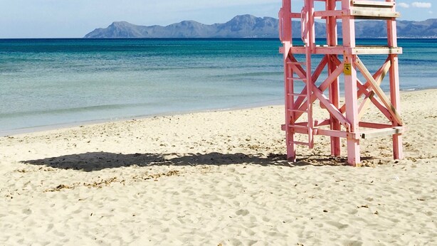 A pink lifeguard tower on a sandy beach with clear blue water and distant mountains.