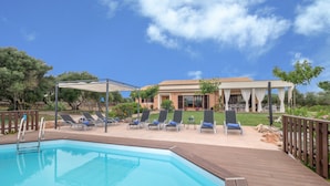 A pool area with a wooden deck, lounge chairs, and a building with a tiled roof in the background.