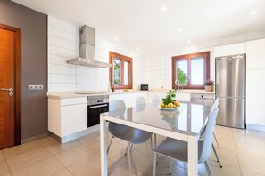 A modern kitchen with white cabinetry, stainless steel appliances, a dining table with chairs, and a window with a view of greenery.